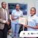 Madam Carolyn Vuni, Manager Technical Monitoring at UCC (M) Awarding a certificate of Participation to one of the Participants alongside Dr. Frank Pio Kiyingi (L).